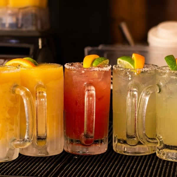 Four colorful margaritas, two yellow, one red, and one light green, garnished with citrus slices.
