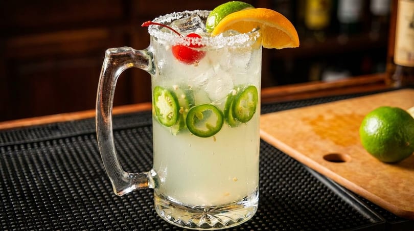 Spicy margarita in a glass mug with jalapeños, lime, orange, and a cherry on a bar.